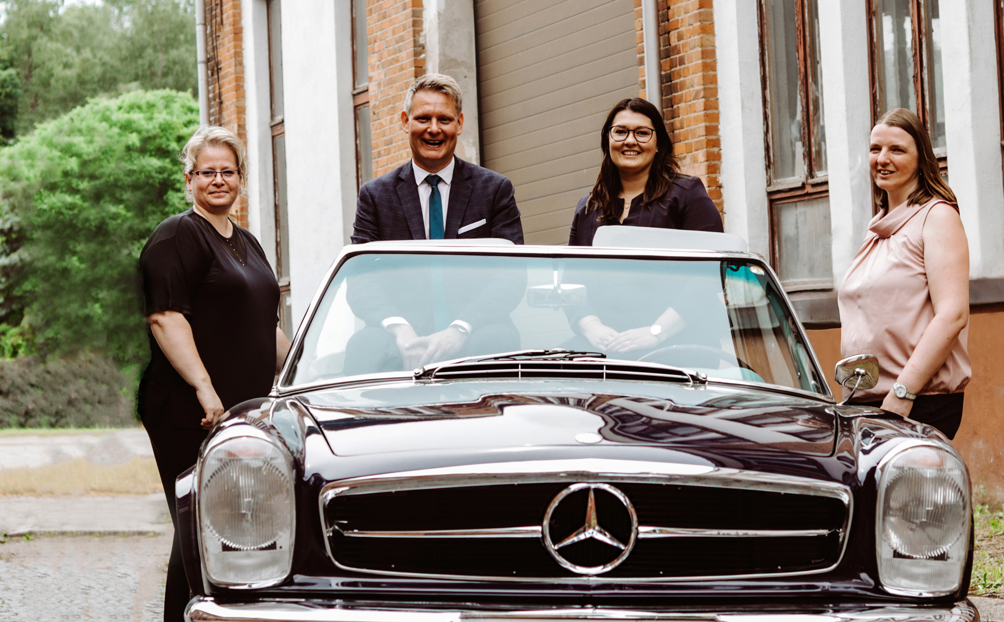 Das Kanzleiteam steht lächelnd um ein klassisches schwarzes Mercedes-Cabriolet. Zwei Personen sitzen im Auto, zwei stehen außen. Die Szene spielt im Freien vor einem alten Backsteingebäude mit Fenstern.